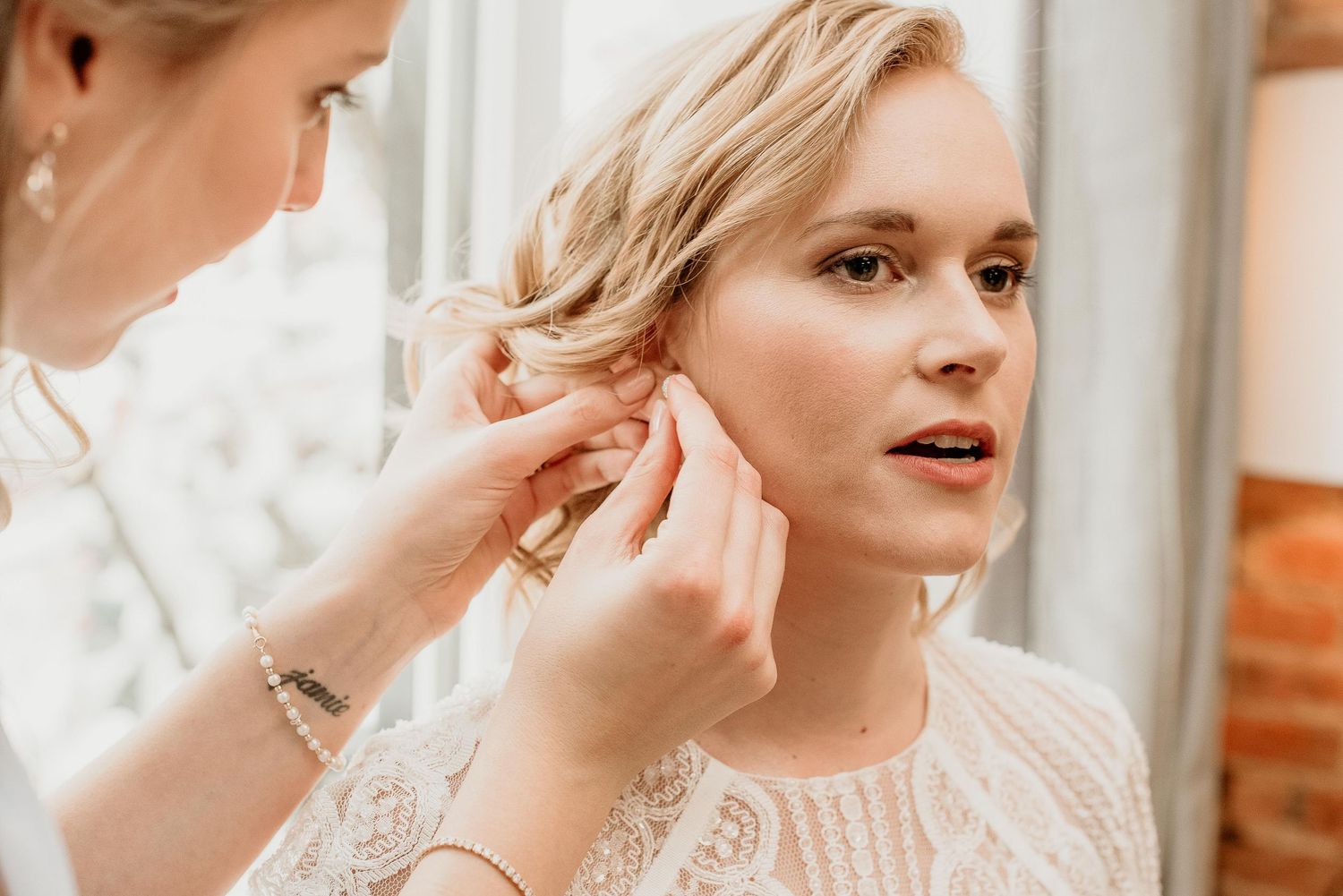 Bridesmaid helping blonde bride put on earrings while getting ready for wedding ceremony.