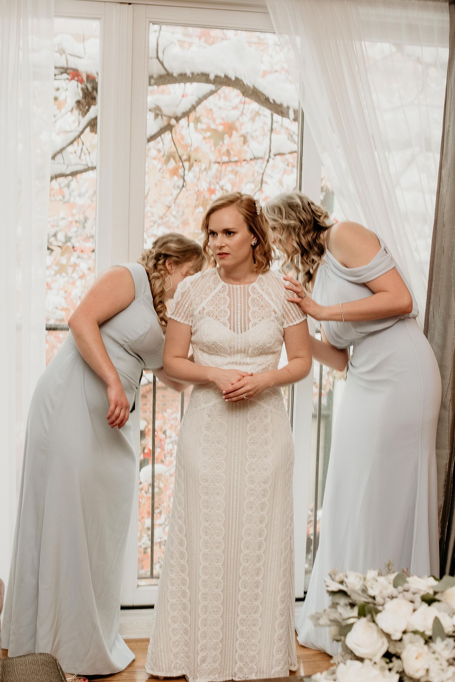 Bride in lace gown assisted by two bridesmaids in silver dresses near a window with floral backdrop.