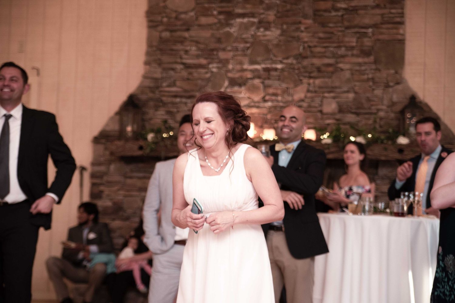 Woman in white dress smiling at wedding reception with guests and stone fireplace in background.
