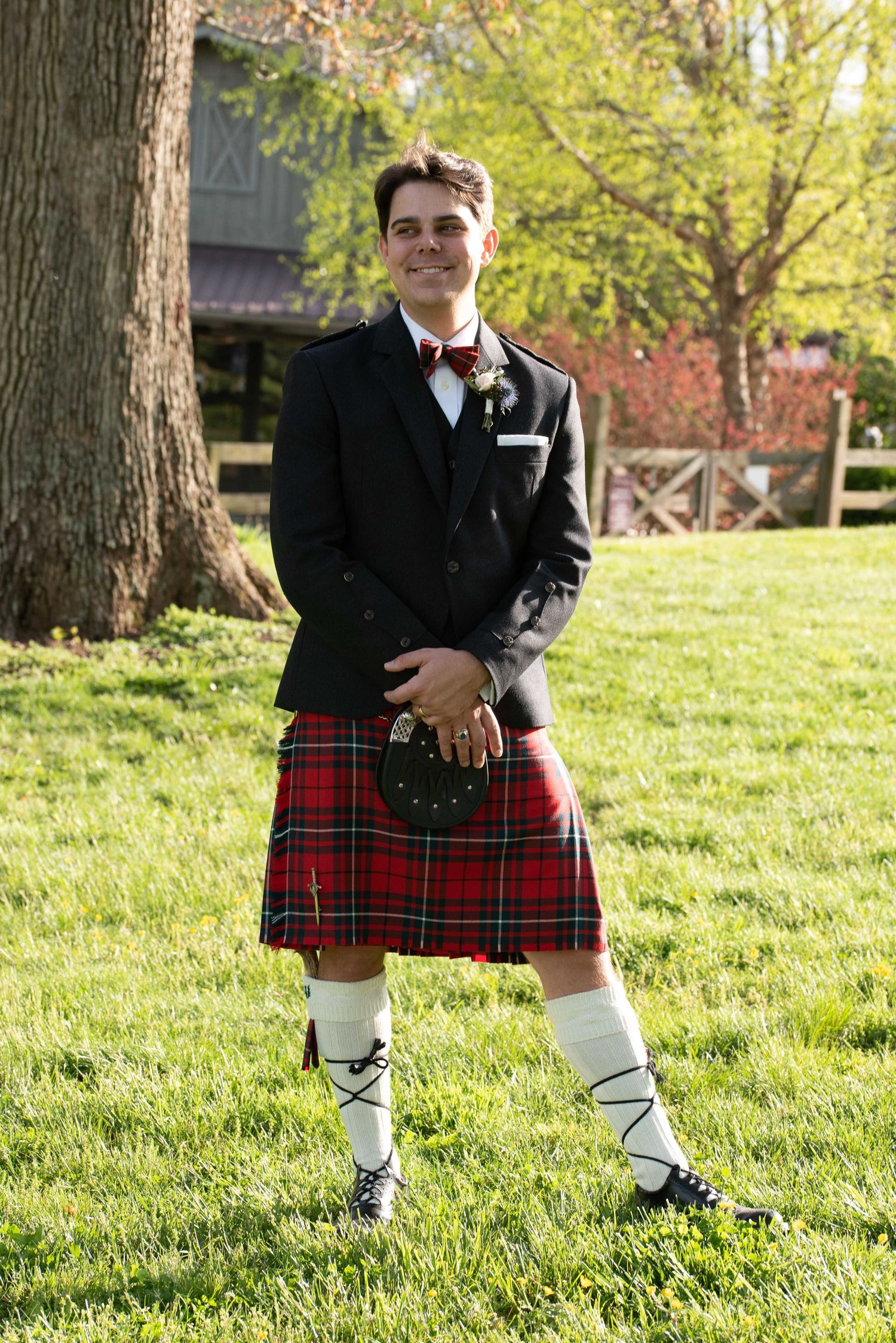 Man in traditional Scottish kilt outfit with black jacket, red tartan, sporran, and knee-high socks standing on green grass.