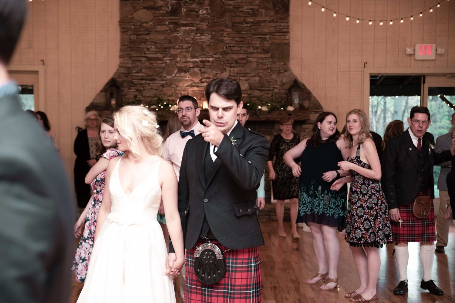 Bride and groom in Scottish tartan kilt holding hands on wedding reception dance floor with guests.