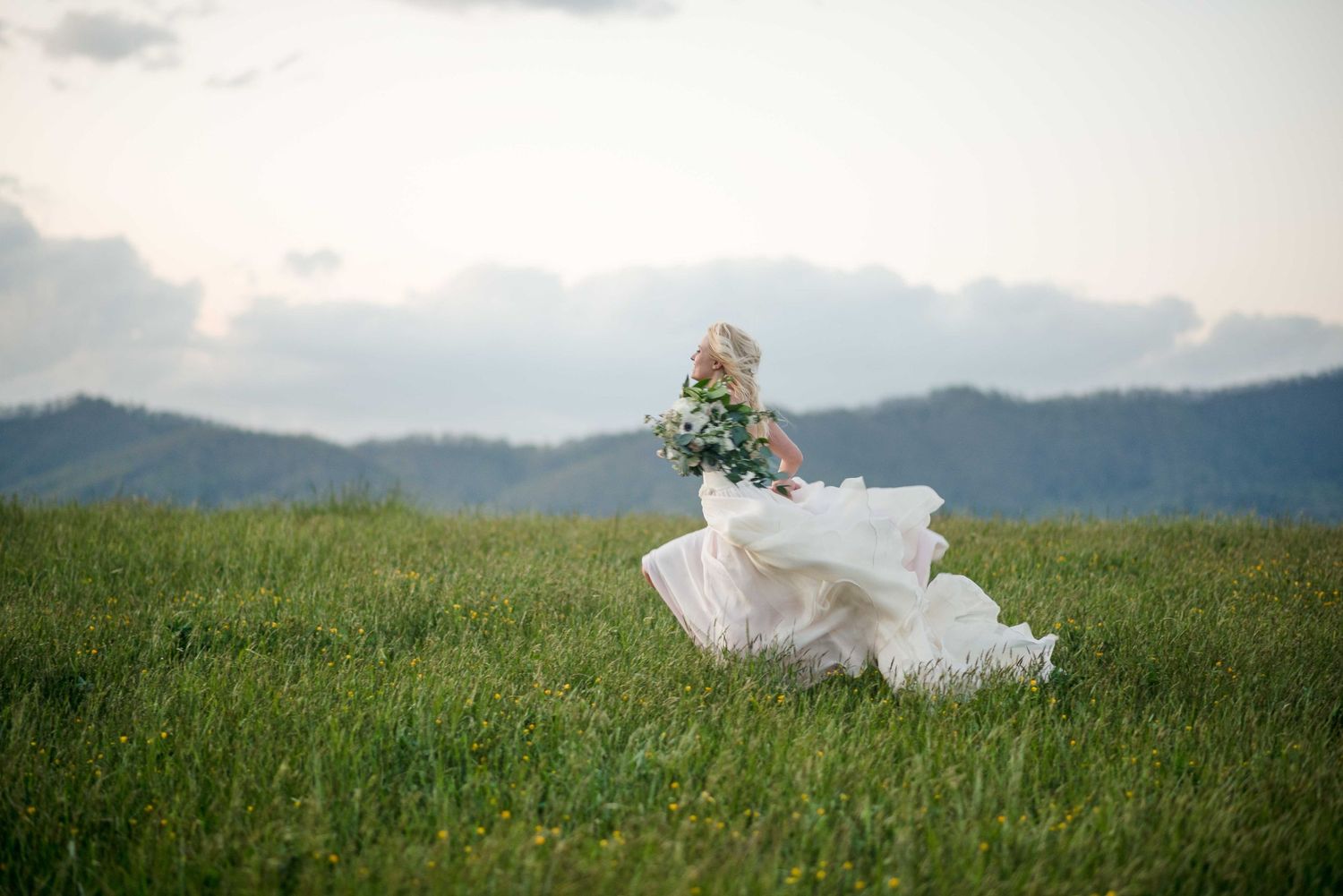 Bride in flowing white wedding gown twirling in lush green field with mountain backdrop on cloudy day.