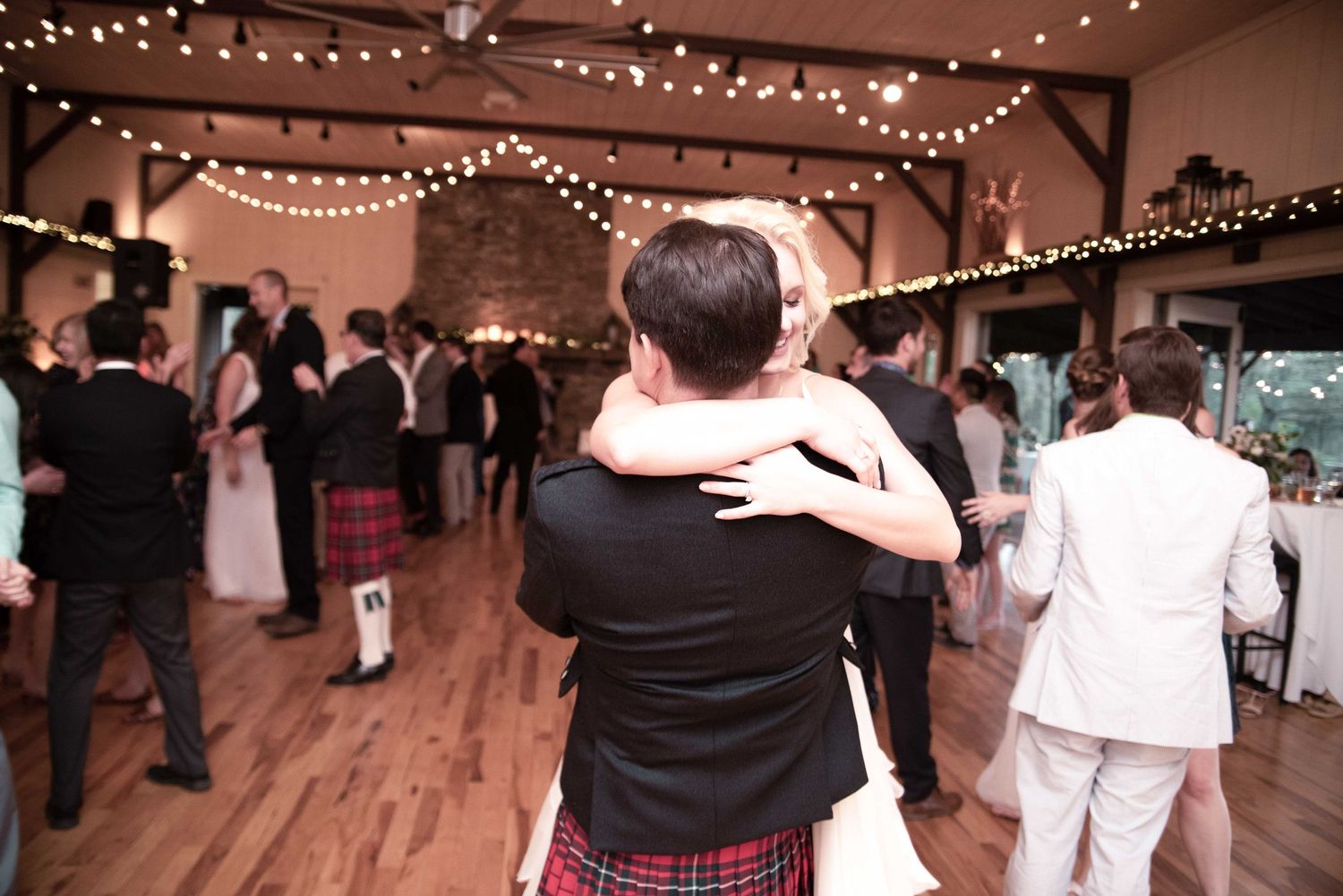 Couple slow dancing at a rustic wedding reception with string lights and guests in background.