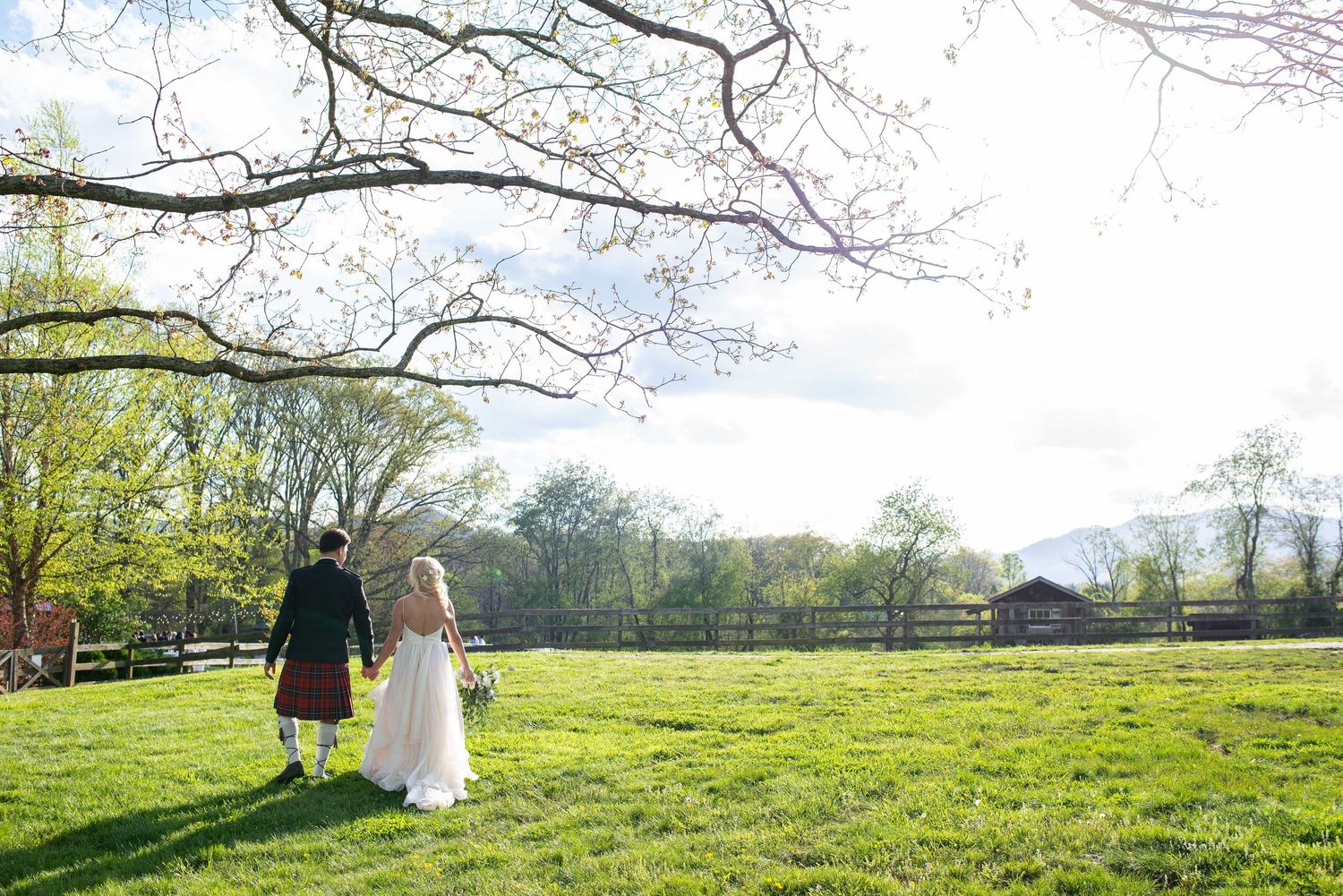 Bride and groom walking hand in hand across lush green meadow at rustic outdoor wedding venue on sunny spring day.