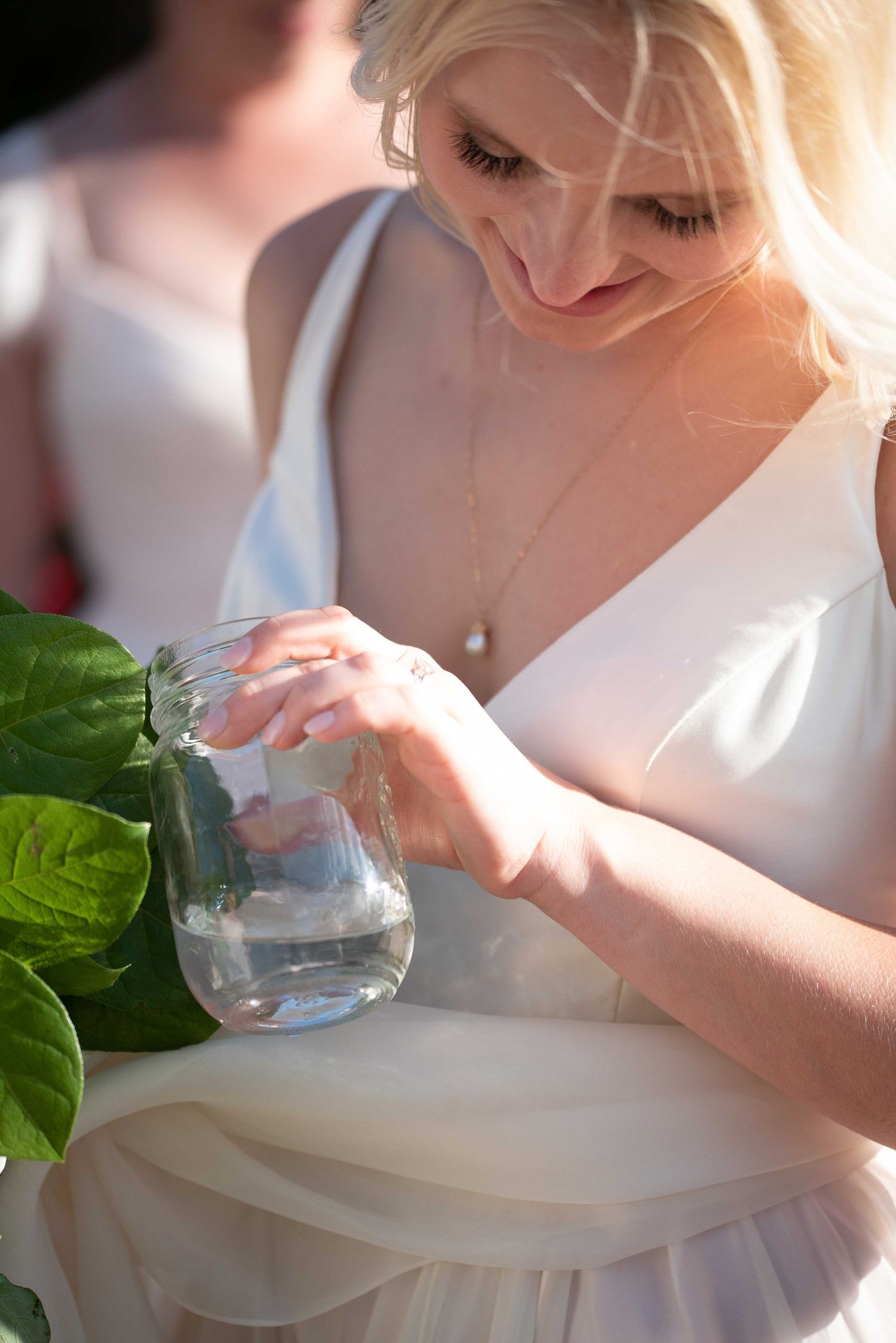 Blonde bride in white dress pouring water into a mason jar, wearing a delicate necklace outdoors.