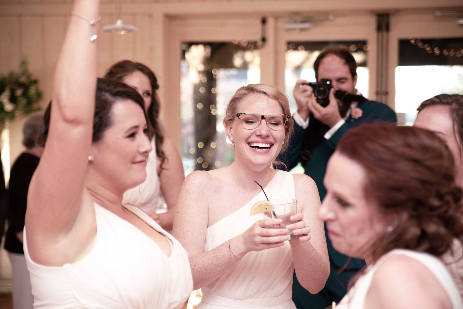 Bridesmaids laughing and celebrating at a wedding reception, wearing white dresses, with guests and photographer in background.