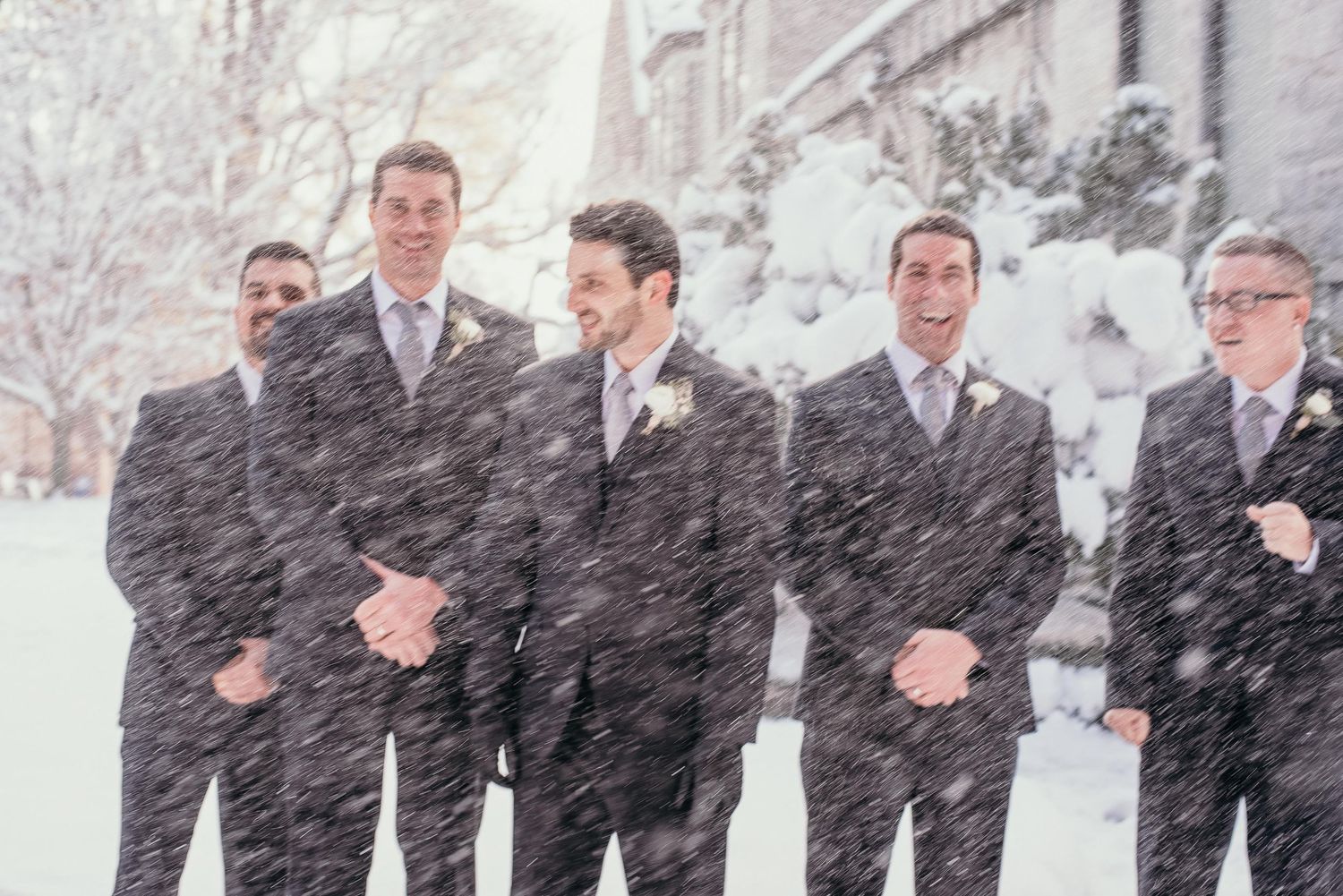 Groomsmen in gray suits laughing together in heavy snowfall during a winter wedding outdoor photo.