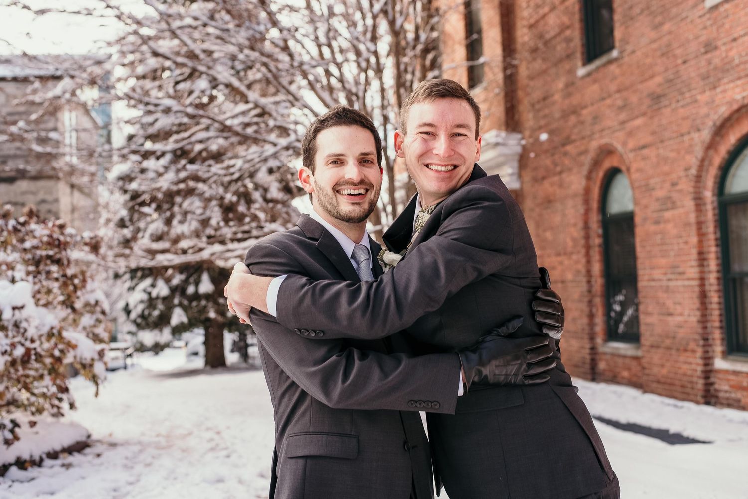 Two men in suits embracing and smiling outdoors in a snowy winter setting near a brick building.