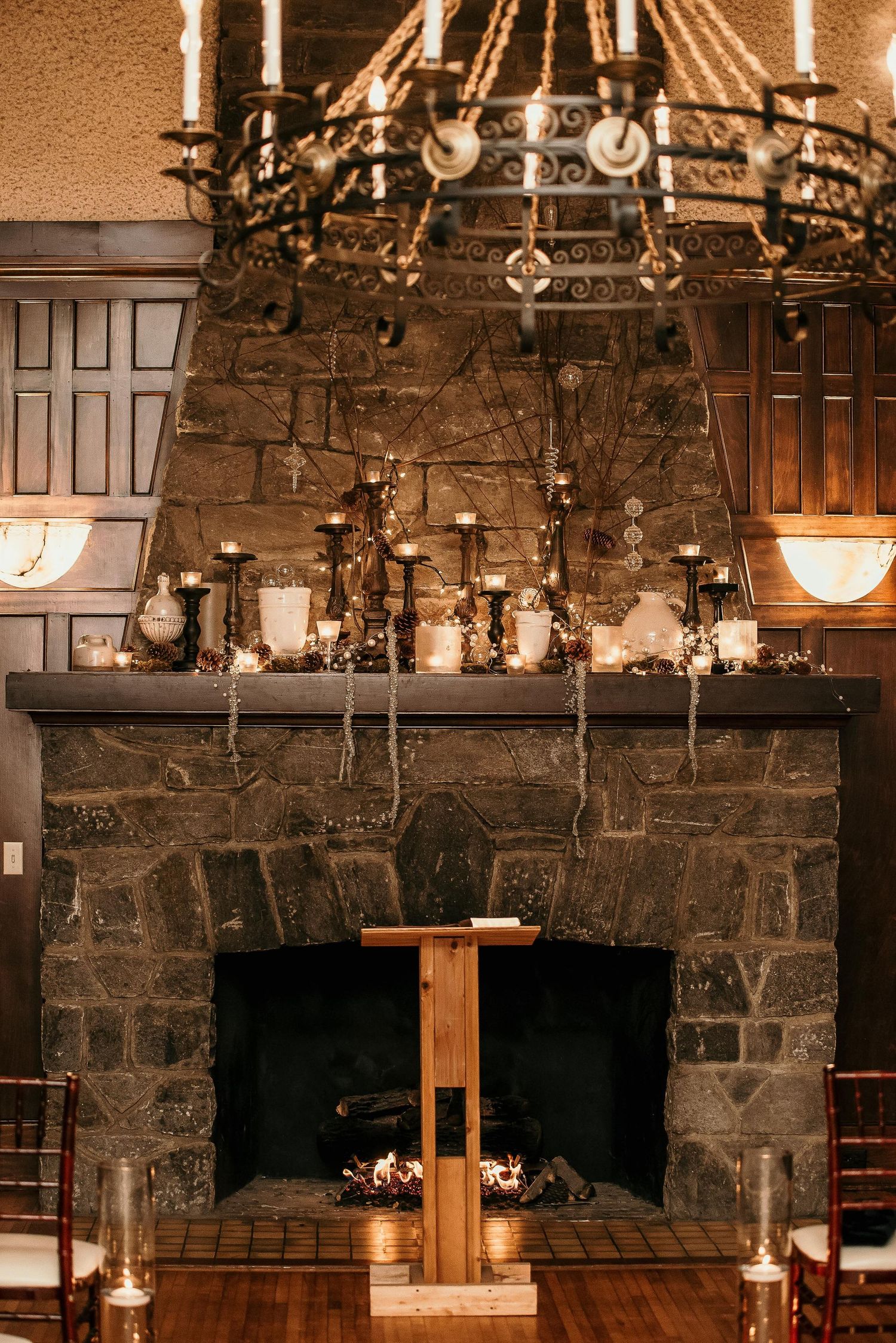 Stone fireplace wedding ceremony setup with candlelit mantel, wooden altar, and elegant chandelier overhead.