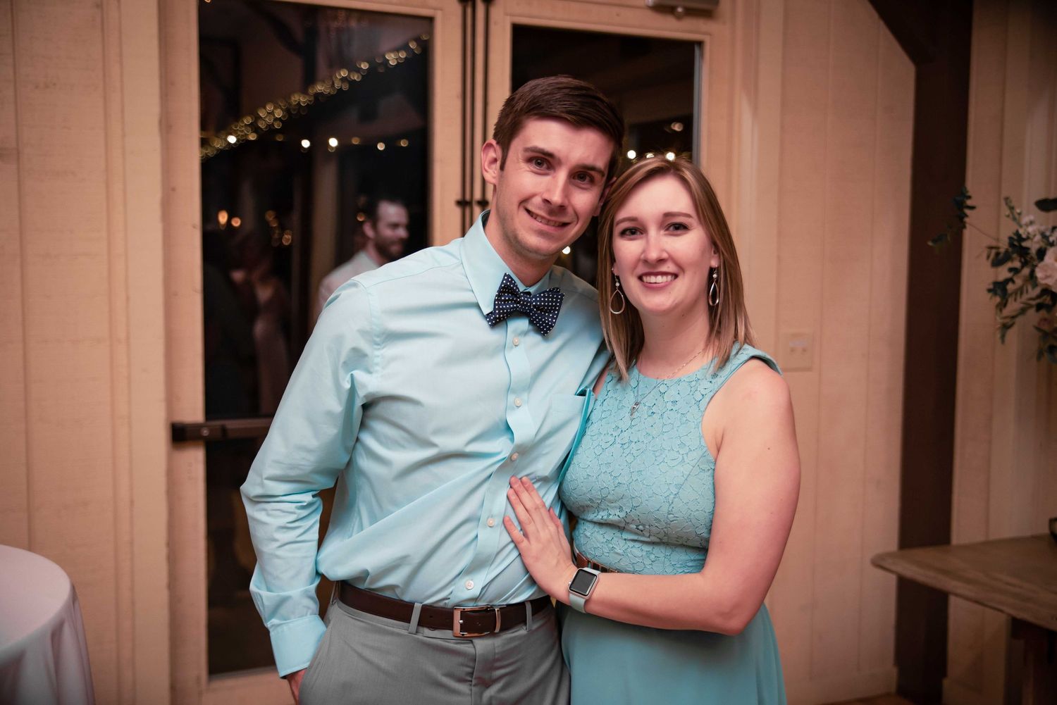 A smiling couple dressed in coordinating teal outfits poses together at an elegant indoor wedding reception.