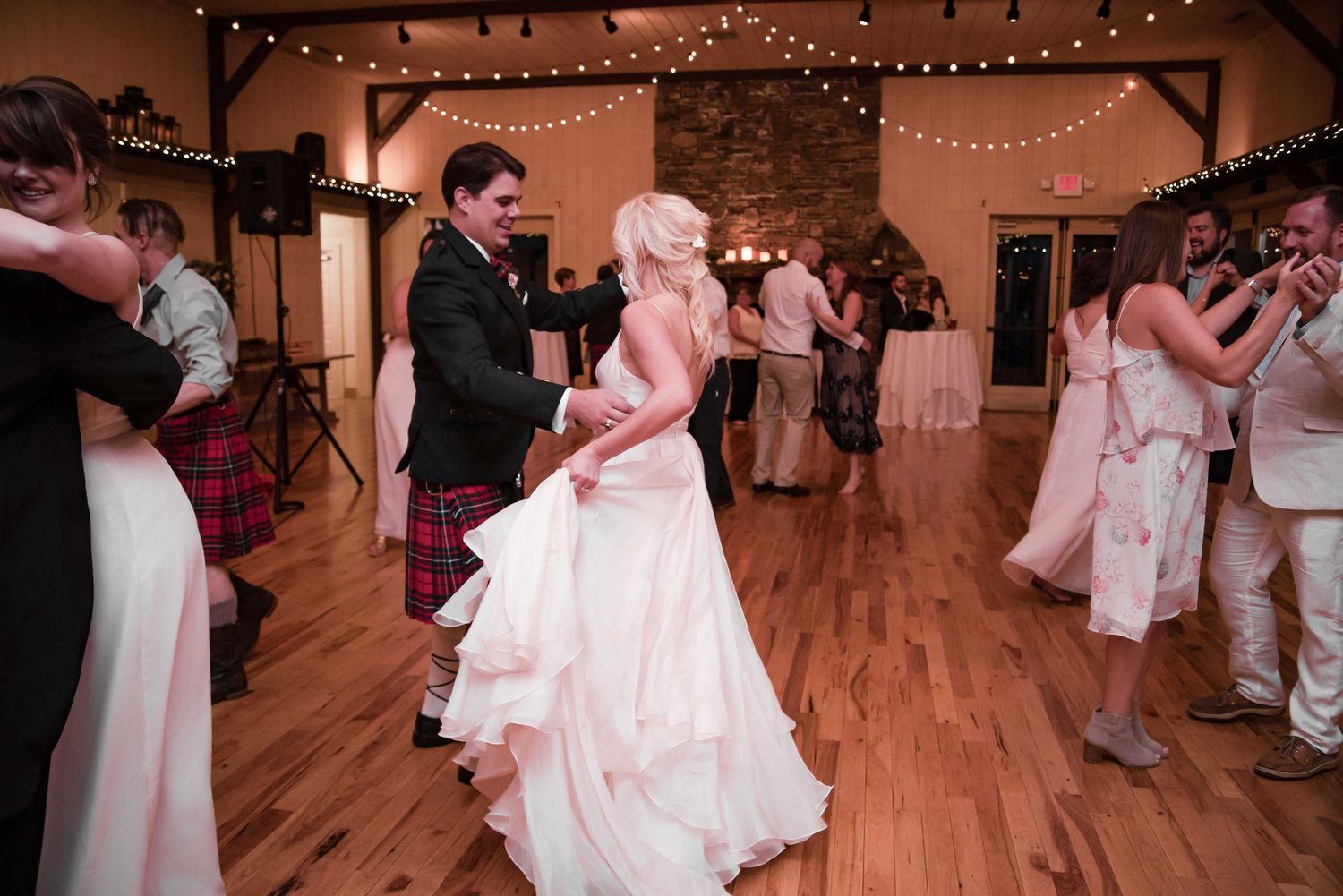 Bride and groom share a first dance at rustic wedding reception venue with string lights and guests dancing.