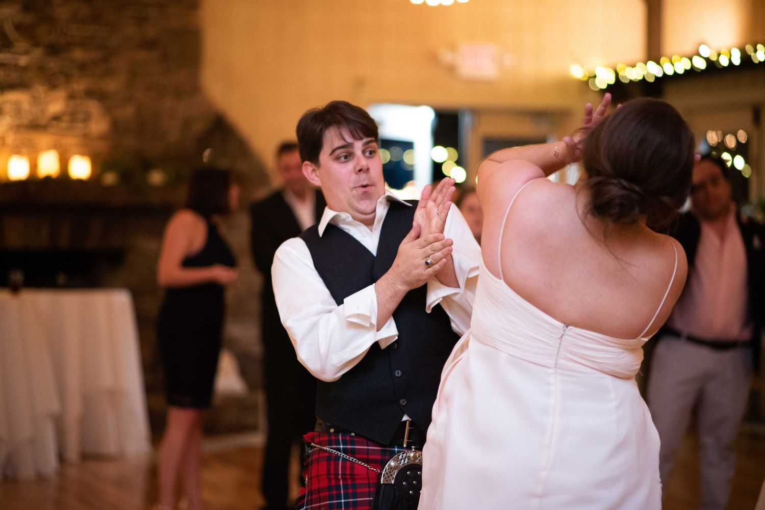 Groom in tartan kilt dancing with bride in white strapless gown at elegant wedding reception.