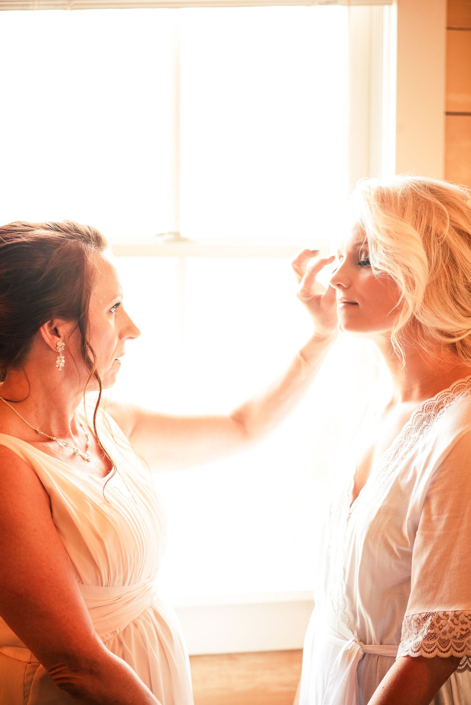 Bride getting makeup applied by bridesmaid near bright window in white lace robe before wedding ceremony.