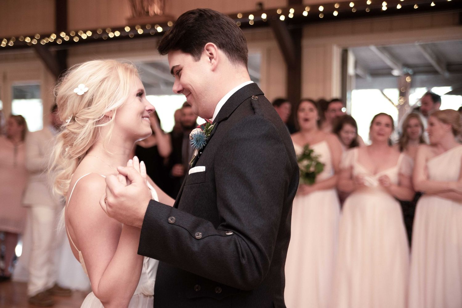 Bride and groom share first dance at wedding reception with bridesmaids in pink dresses cheering in background.