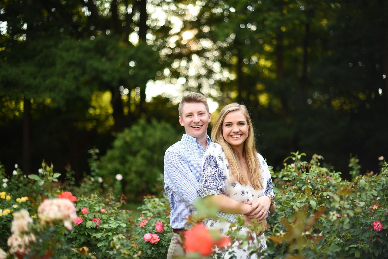 Couple smiling together in a lush rose garden with green trees in the background during golden hour.