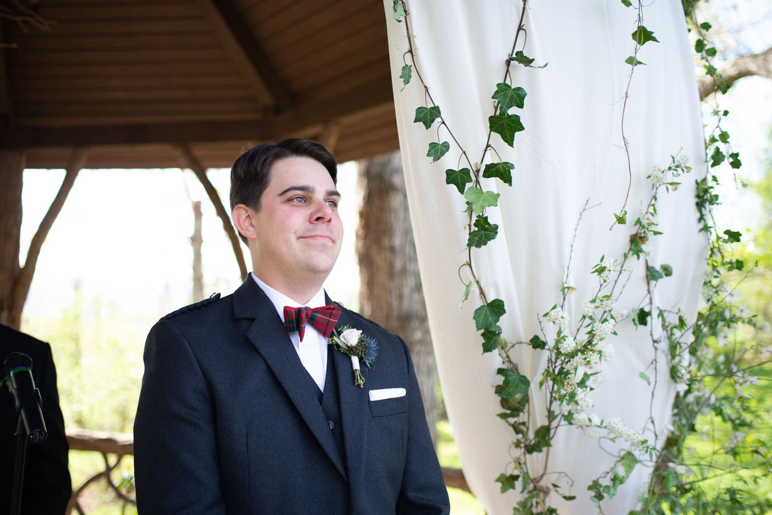 Groom in a dark suit with burgundy bow tie smiling emotionally at a wedding ceremony beneath a floral arch.
