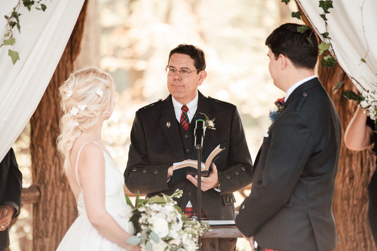 Officiant reads from a book during an outdoor wedding ceremony with bride and groom under a floral arch.