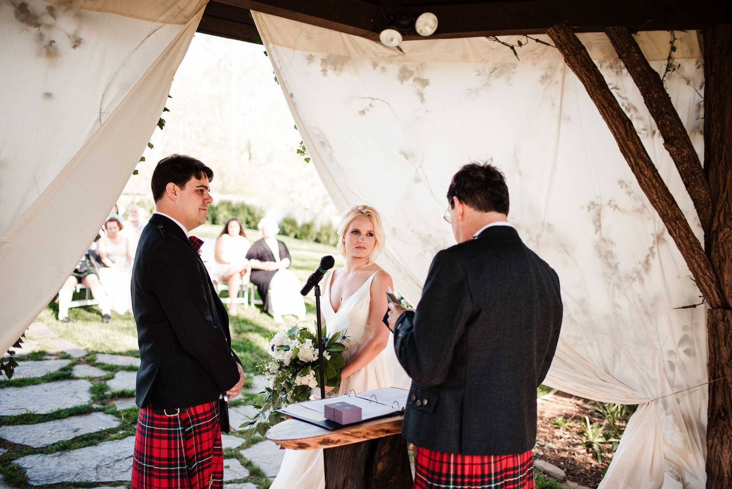 Outdoor wedding ceremony under a draped canopy with bride, groom, and officiant wearing traditional Scottish kilts.