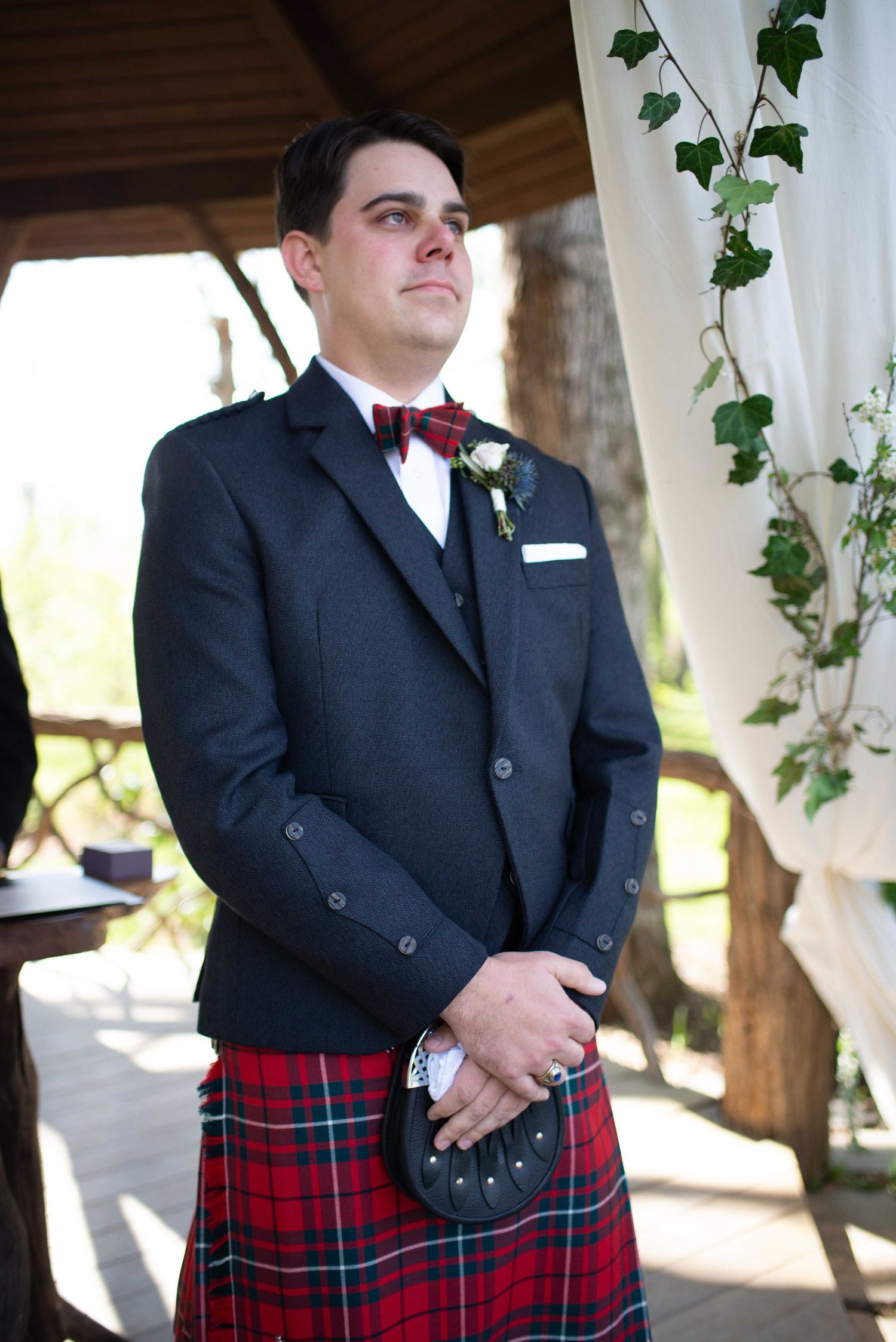 Groom in traditional Scottish tartan kilt and dark jacket with red bow tie standing at outdoor wedding ceremony.