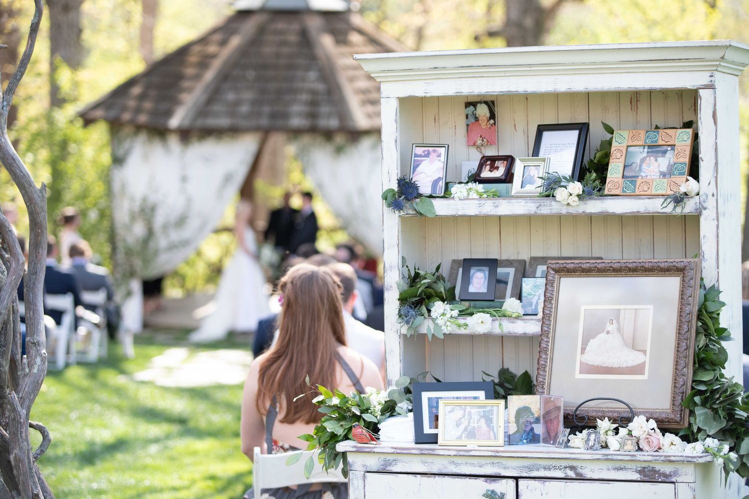 Outdoor wedding ceremony with a vintage dresser displaying framed family photos and floral arrangements near a gazebo.