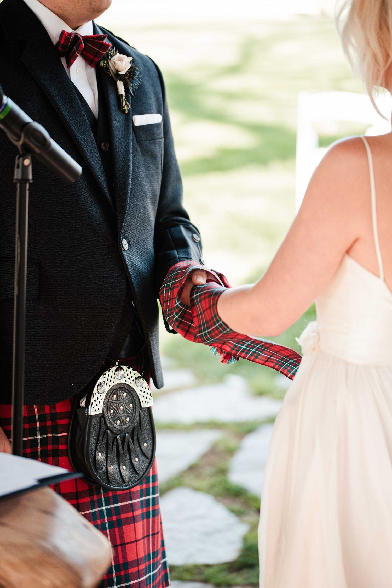 Groom in red Royal Stewart tartan kilt and sporran holds hands with bride in white dress at outdoor wedding ceremony.