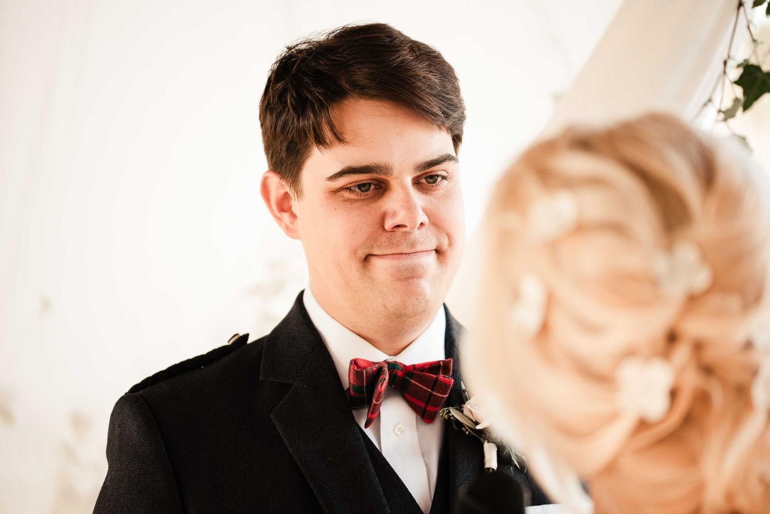 Groom in black suit and plaid bow tie smiling at bride during wedding ceremony.