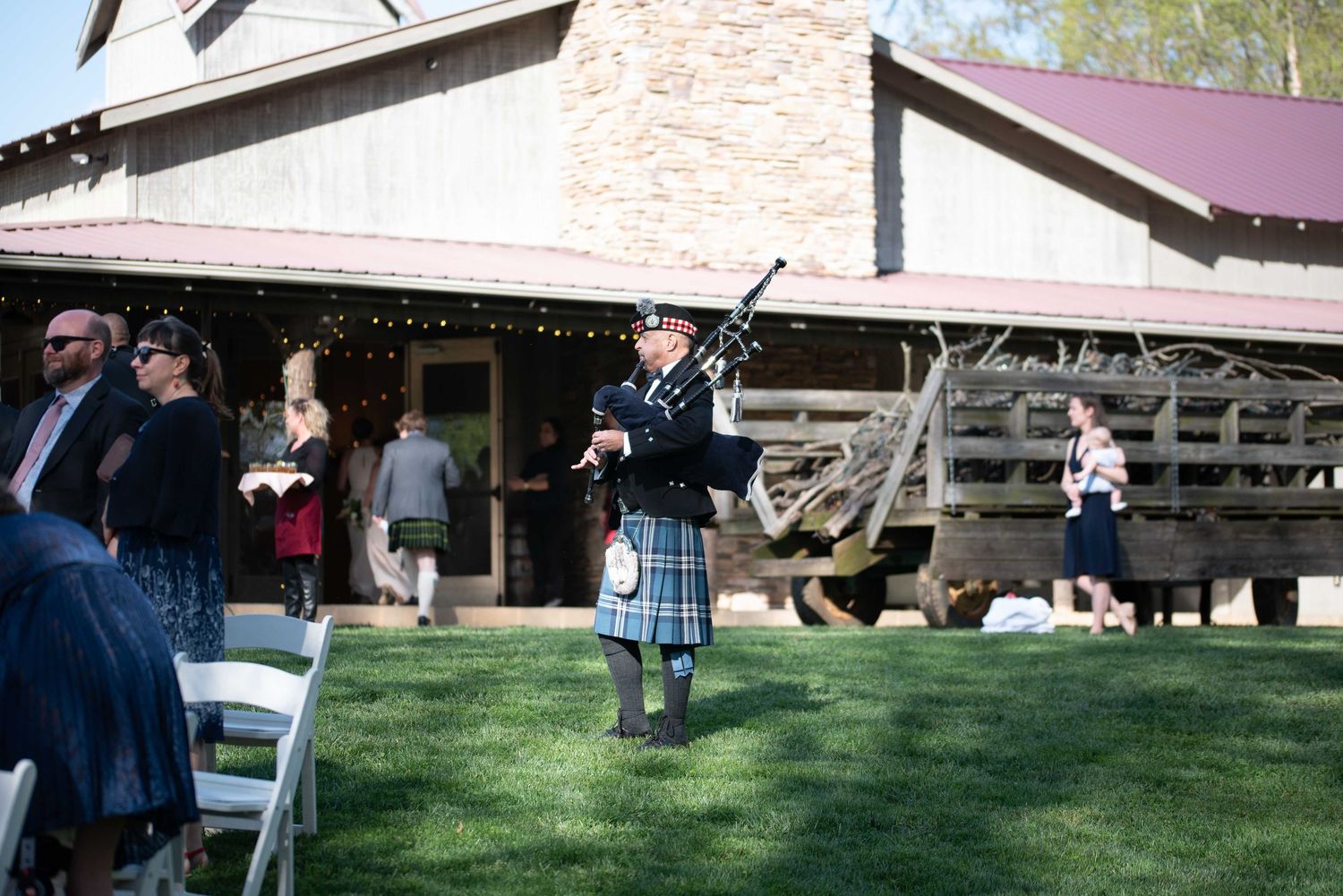 Bagpiper in blue tartan kilt performing outdoors at a rustic barn wedding venue on a sunny day.
