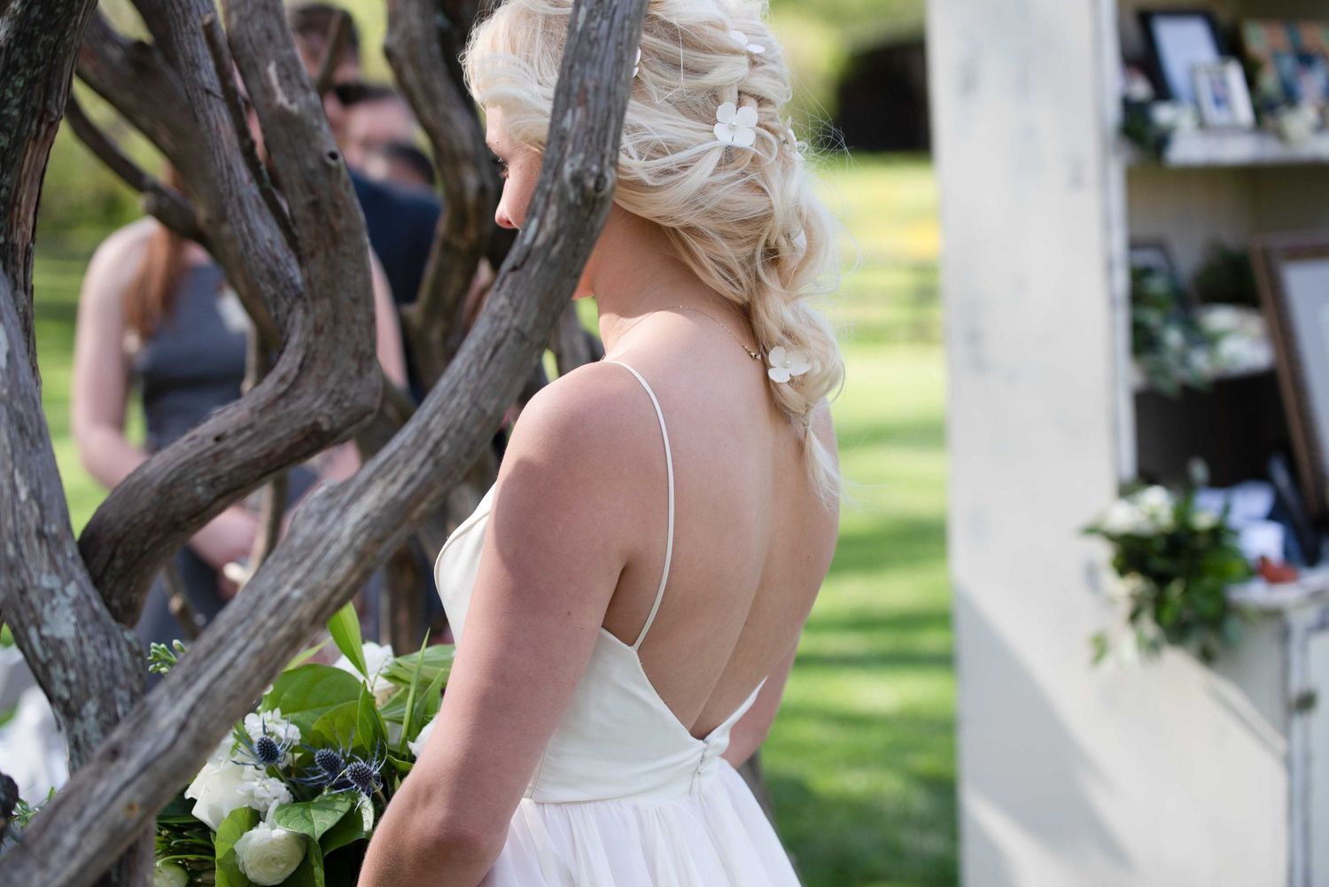 Bride in backless white gown holds bouquet beside rustic branch arch at outdoor wedding.
