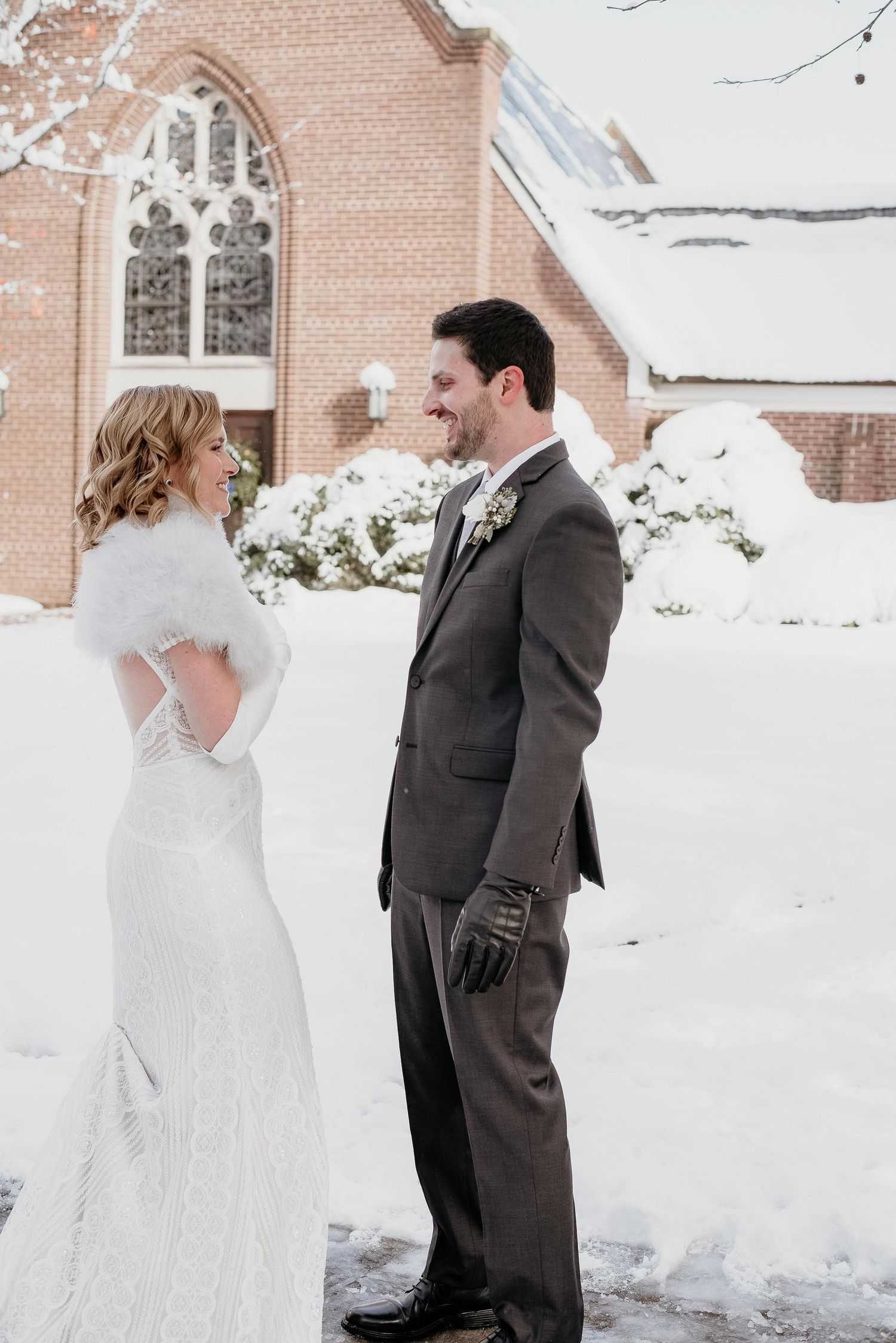 Bride and groom share a first look moment outside a snow-covered brick church in winter wedding attire.