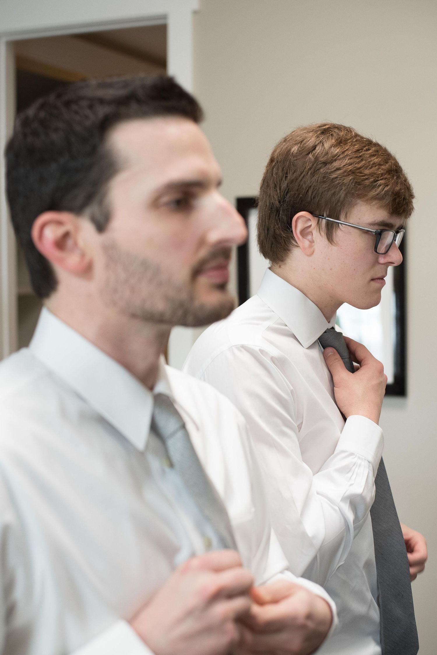Two men in white dress shirts adjusting their ties while getting ready for a formal event.