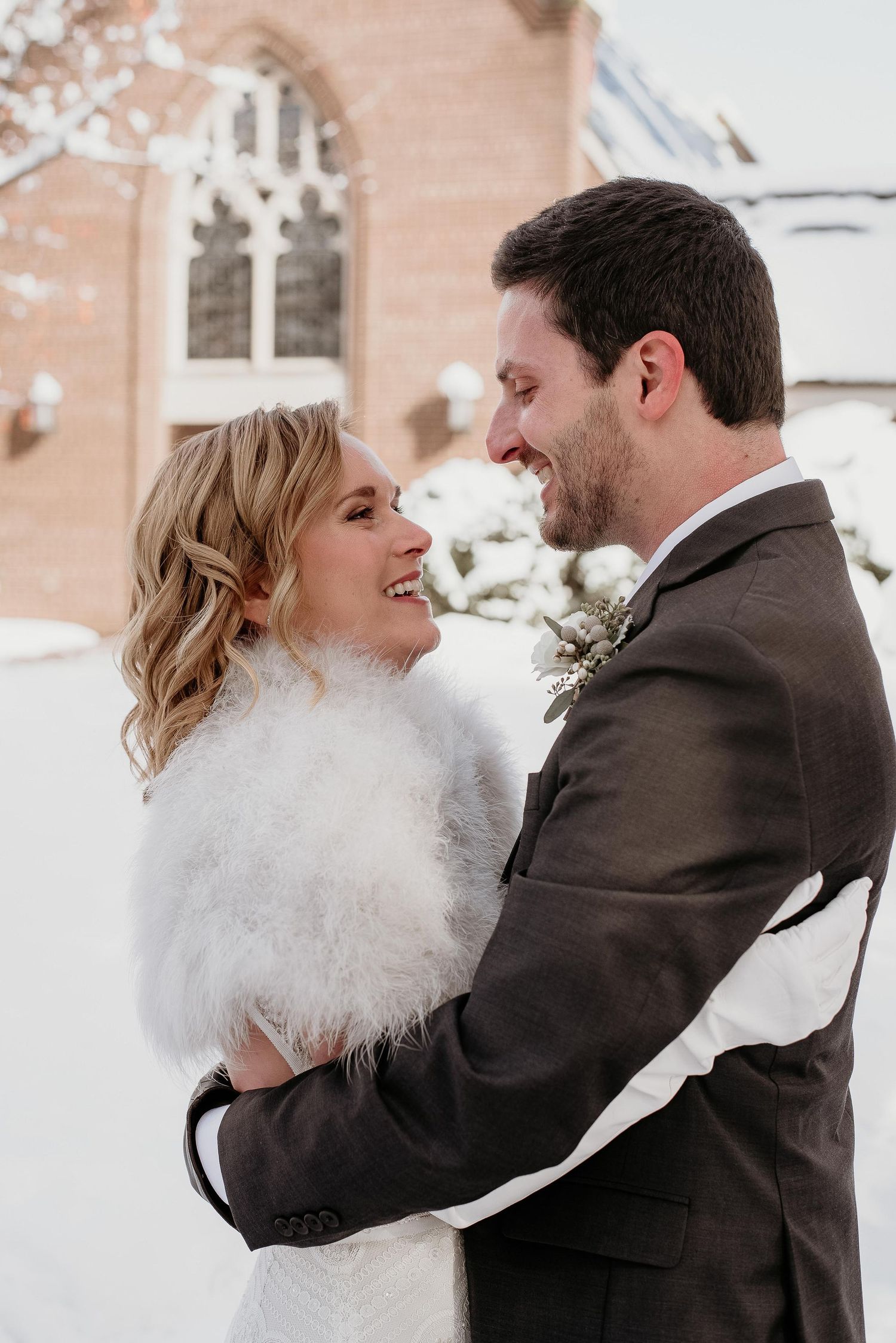 A joyful couple embraces in the snow outside a church, bride wearing a white fur stole and groom in a gray suit.