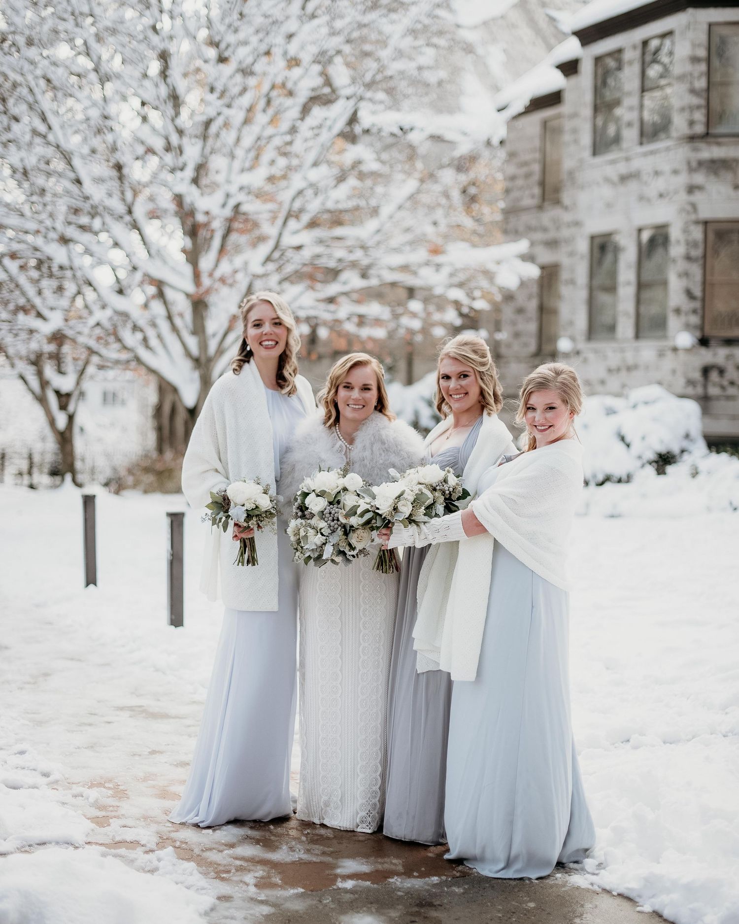 Bride in white lace gown with three bridesmaids in light blue dresses holding white bouquets in a snowy winter setting.