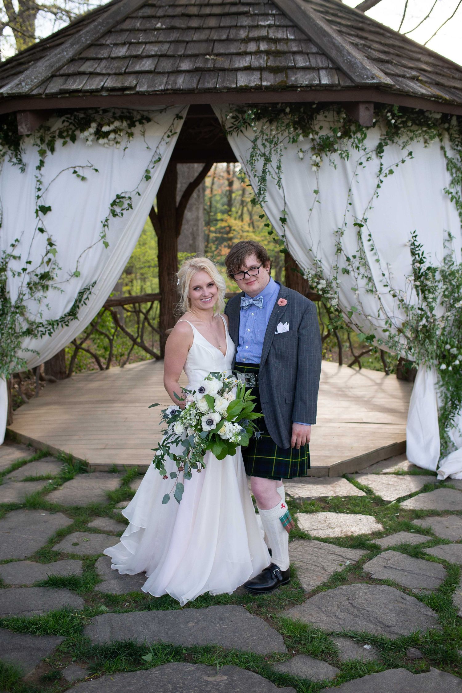 Bride in white gown and groom in tartan kilt pose together under a flower-draped gazebo on their wedding day.