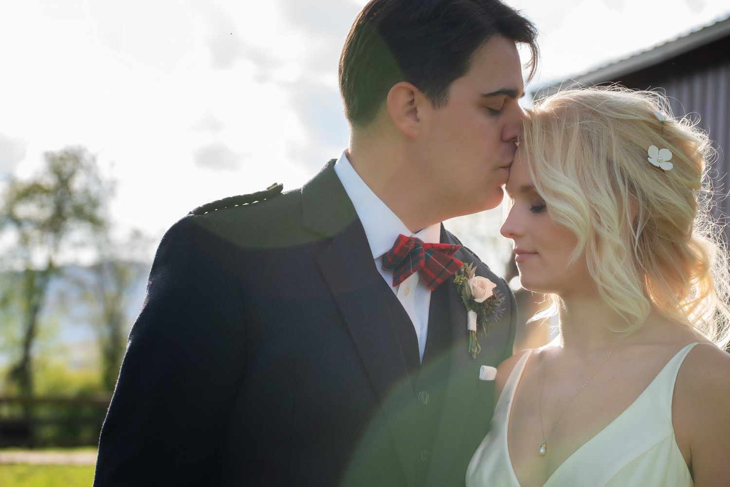 A groom in a navy suit kisses his blonde bride in a white dress outdoors on their wedding day.