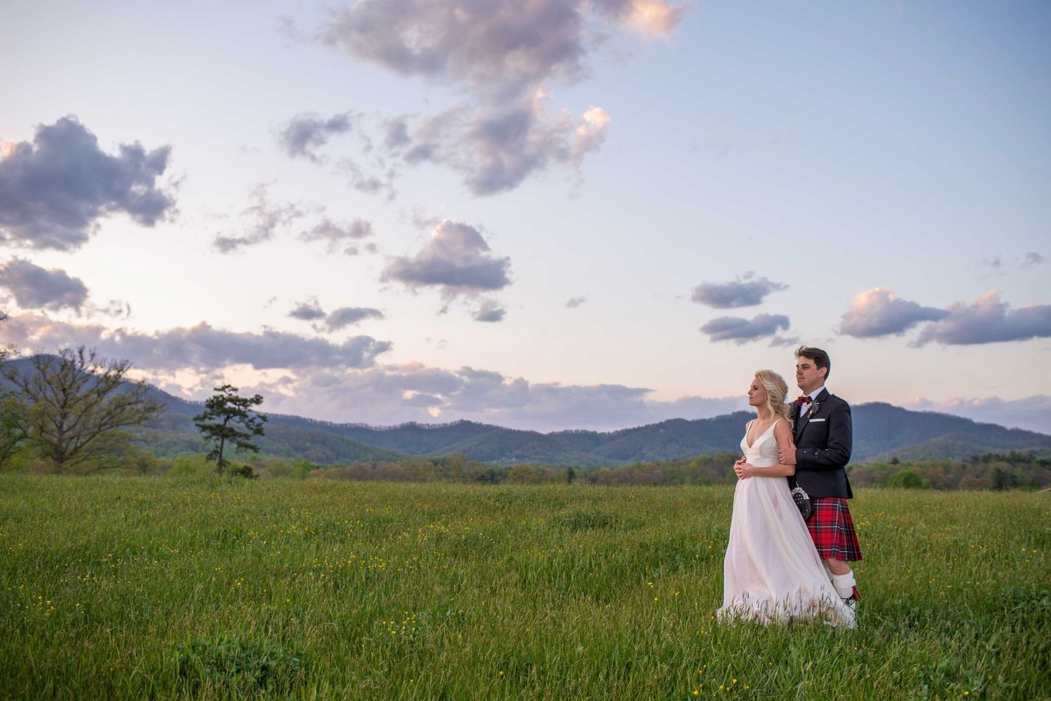 Bride and groom stand in lush green meadow at sunset with mountain backdrop and dramatic clouds.