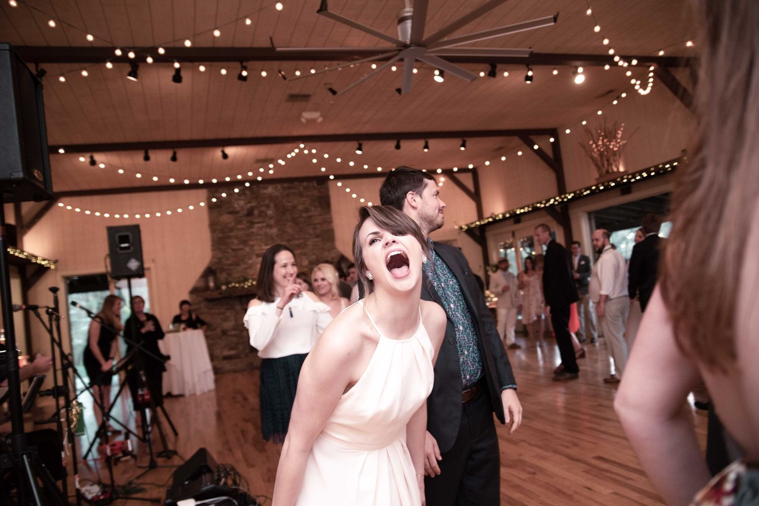 Bride laughing joyfully on wedding reception dance floor with string lights and guests celebrating in rustic venue.