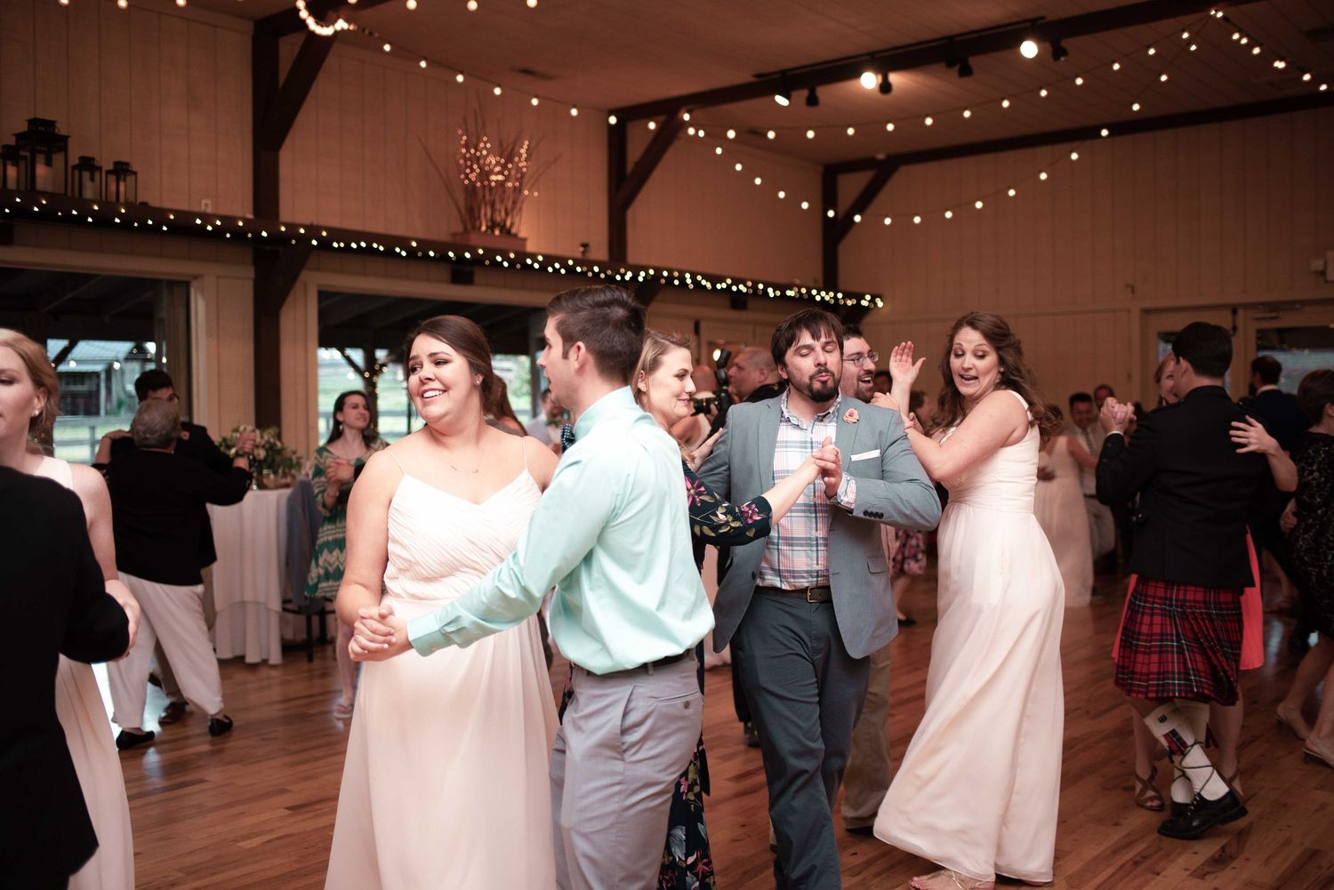 Guests dancing joyfully at a rustic barn wedding reception with string lights and wood-paneled walls.