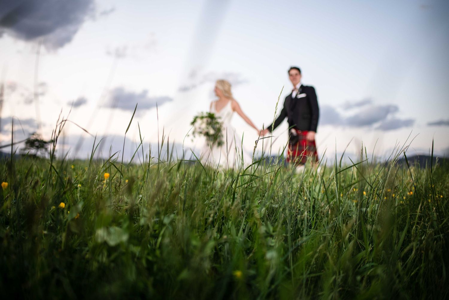Bride and groom walk hand in hand through lush green meadow with dramatic cloudy sky backdrop.