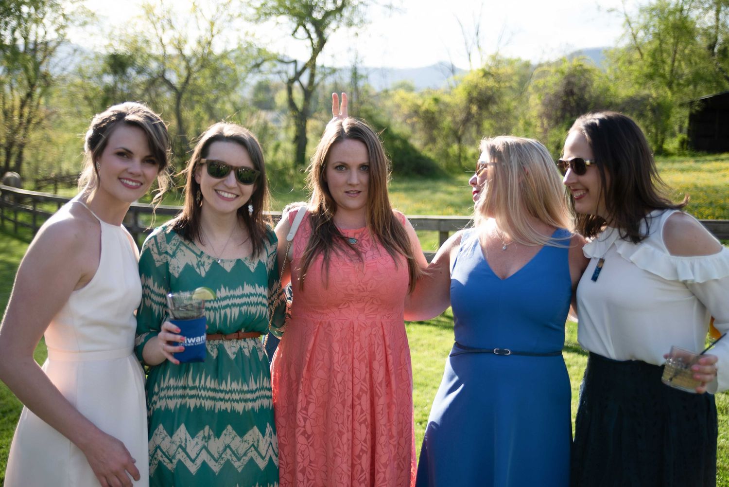 Five women smiling and posing together outdoors at a sunny garden party, wearing colorful summer dresses.