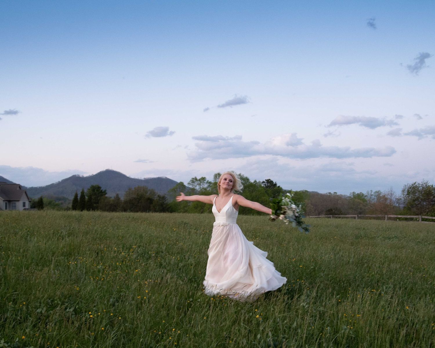 Bride in white gown twirling in green meadow with mountain backdrop at dusk, holding bouquet.