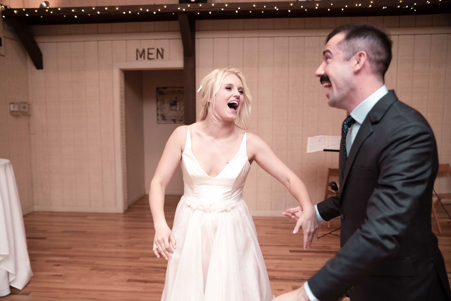 Bride and groom laughing joyfully during wedding reception dance in rustic venue.