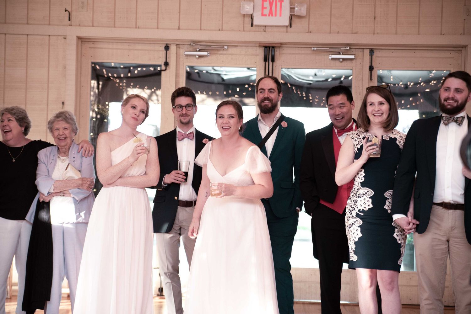 Wedding party poses together smiling in a decorated venue, featuring bride in white gown and guests in formal attire.