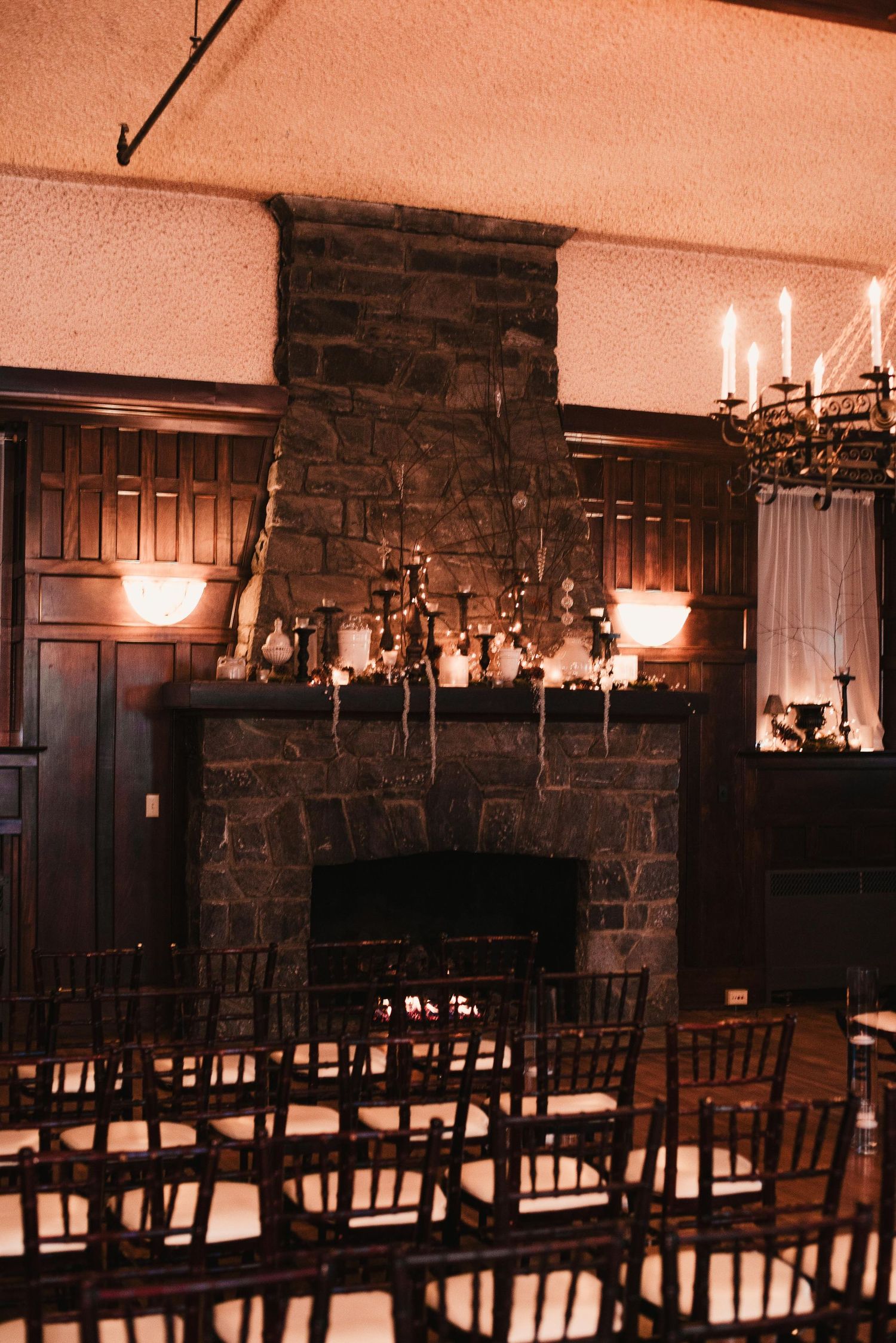 Dark romantic wedding ceremony room with stone fireplace, candelabras, and chiavari chairs in warm candlelight.