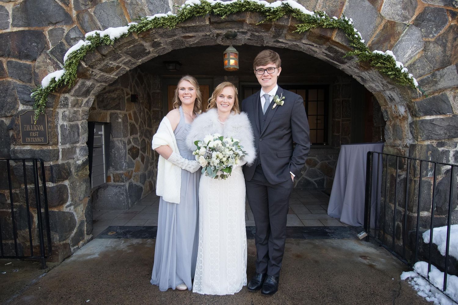 Bride in white gown with bouquet poses with bridesmaid and groomsman under stone arch in winter wedding.