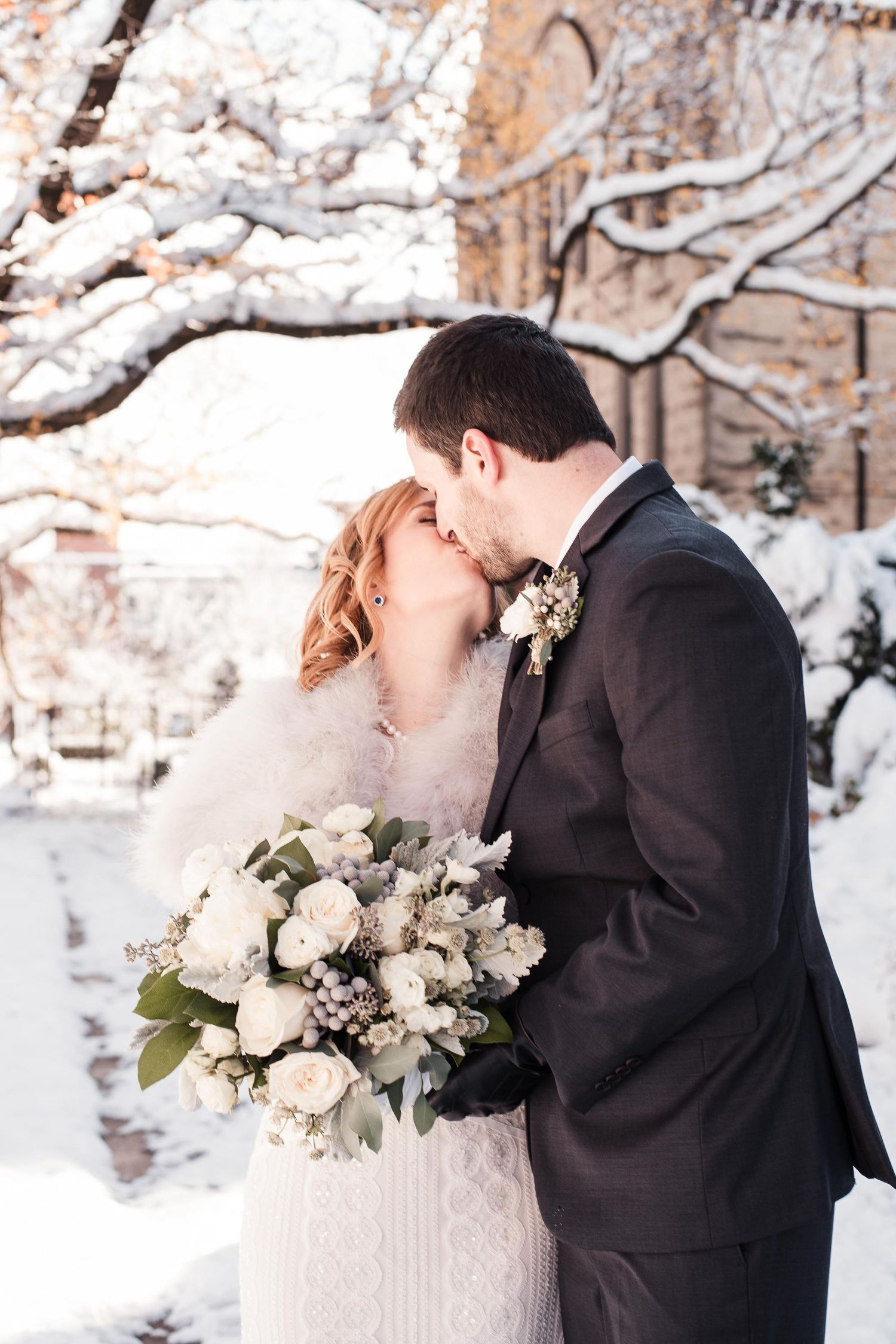 Bride and groom share a kiss in a snowy winter wonderland, bride holding a white rose bouquet.