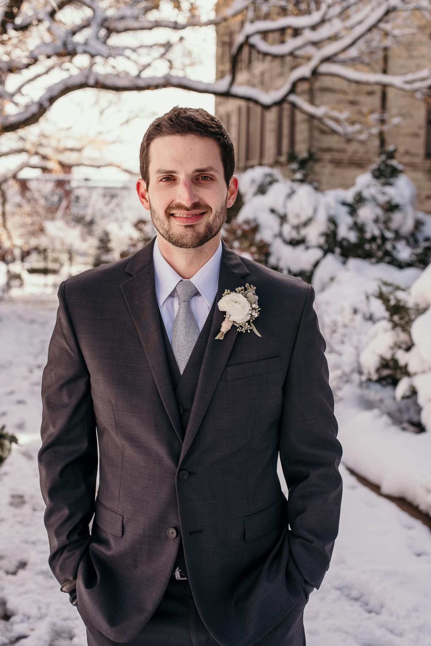 Smiling groom in dark suit with boutonniere poses outdoors in snowy winter garden setting.
