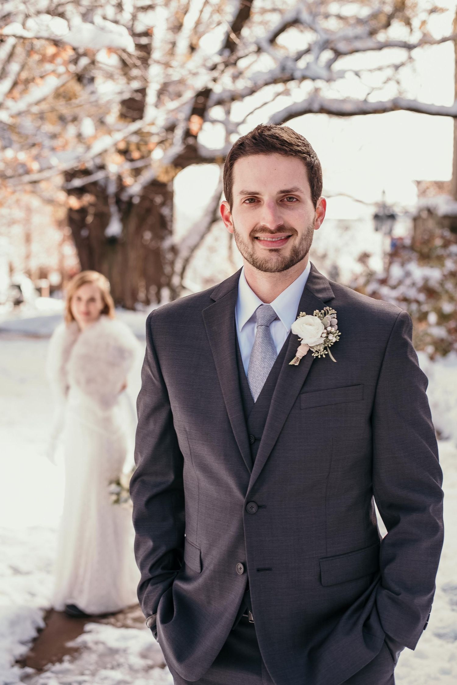 Groom in gray suit with boutonniere smiles outdoors in snow, bride in white dress and fur wrap stands behind him.