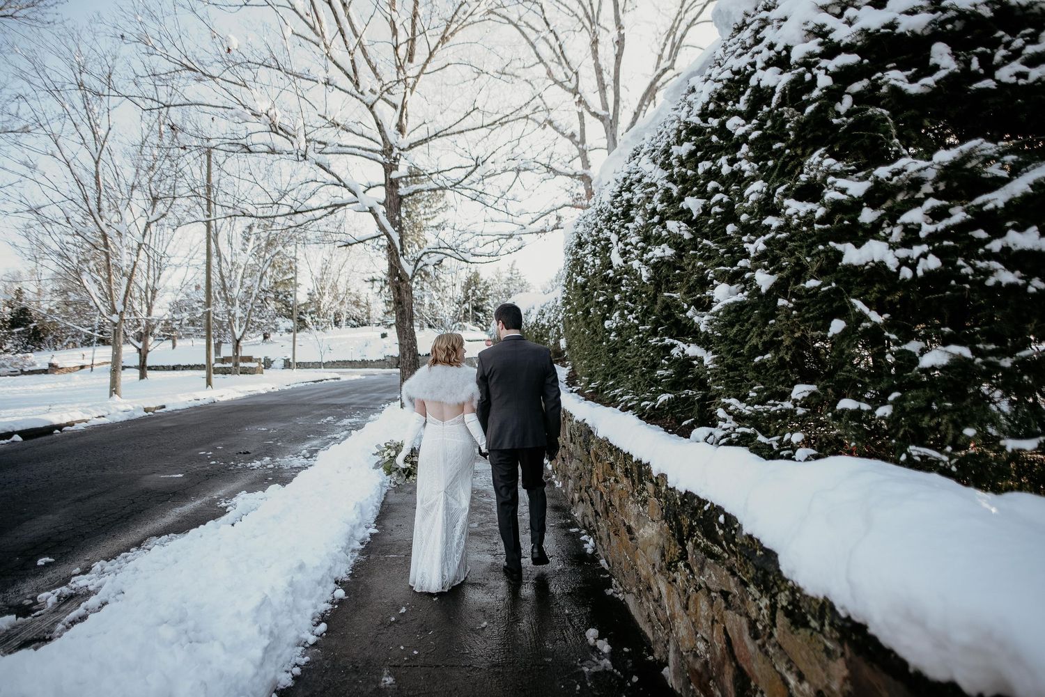 Wedding couple walks hand in hand along a snowy path beside a stone wall on their winter wedding day.