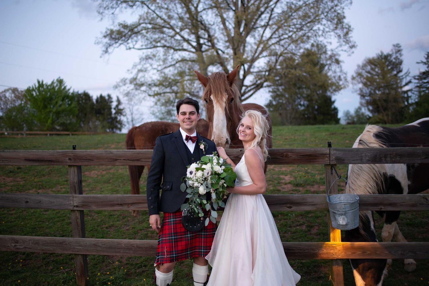 Bride and groom pose by rustic wooden fence with horses on a farm wedding venue, groom wearing traditional Scottish kilt.