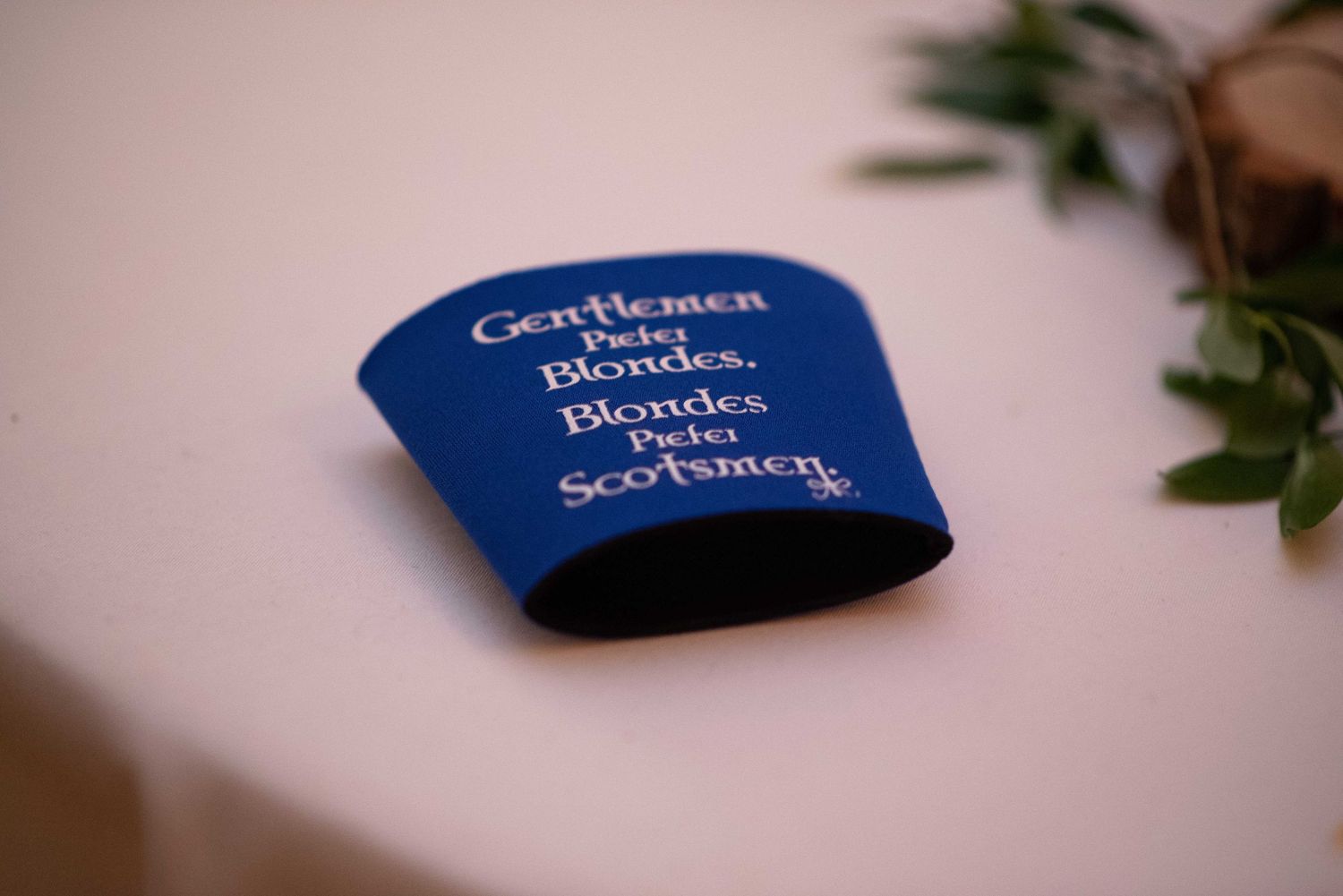 Blue koozie on white table reading Gentlemen Prefer Blondes, Blondes Prefer Scotsmen.